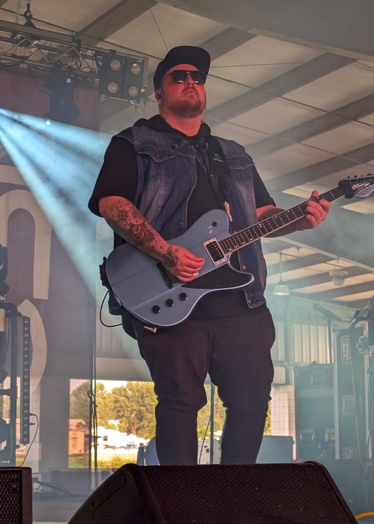 A musician performing on stage with a blue guitar, wearing sunglasses and a denim vest, under stage lighting.