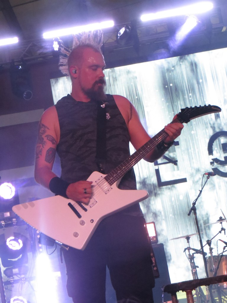 A musician with a mohawk hairstyle playing a white electric guitar on stage, with vibrant lights and a backdrop featuring visual effects.