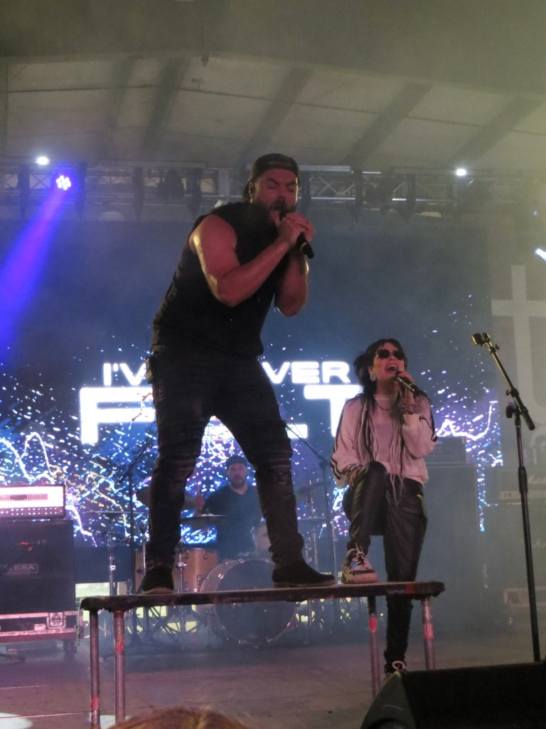 A male singer performing energetically on stage with a microphone, while a female vocalist sits on a platform, both dressed in stylish rocker attire, during a live music event.