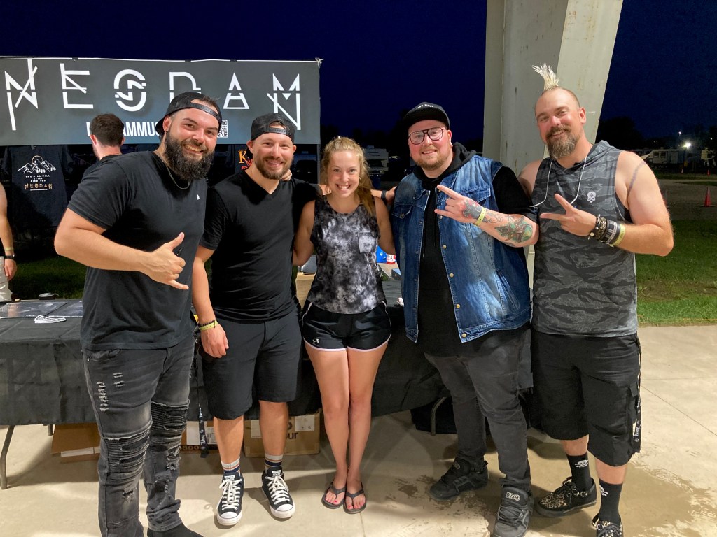 A group photo of the band NESDAM members and a fan, standing in front of a merchandise table at night. The band members are dressed in casual black attire, while one member wears a denim jacket. The background features a sign with the band's name.