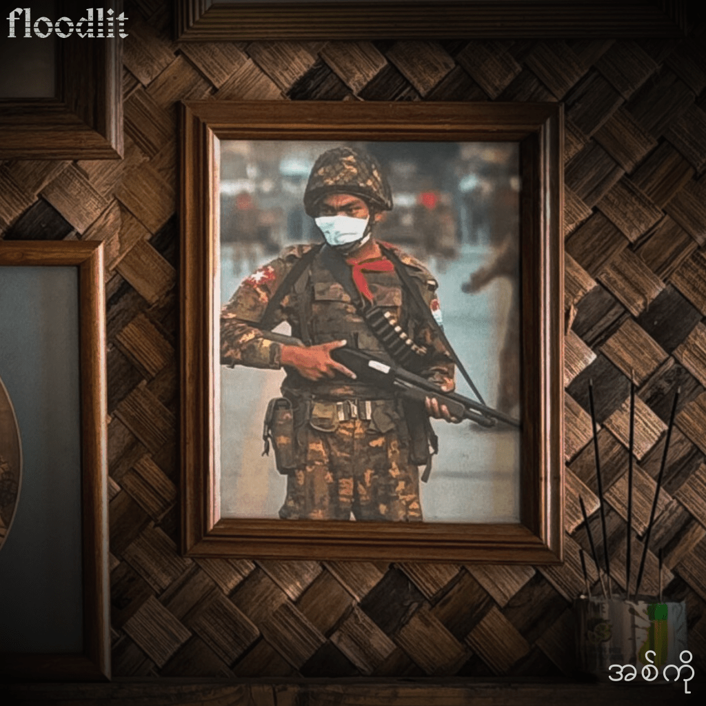 A framed photograph of a soldier wearing a mask and armed with a rifle, standing in a military uniform, displayed on a woven wall.
